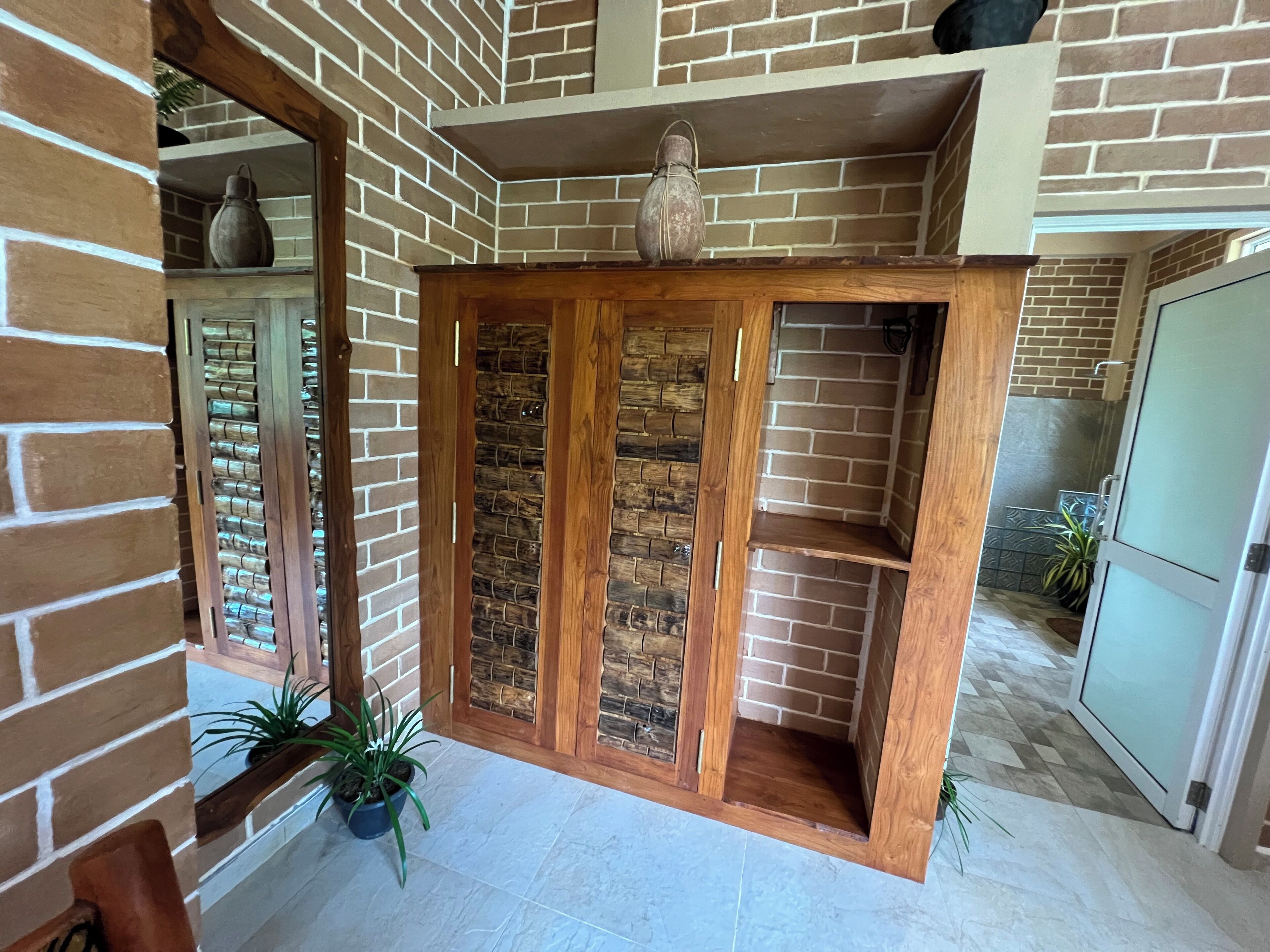 A room with brick walls, wooden doors, and tiled flooring. There are potted plants and a large vase on a shelf.