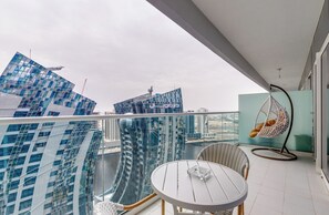 Un balcón con vista a edificios modernos, una silla colgante y una mesa con un recipiente de vidrio.