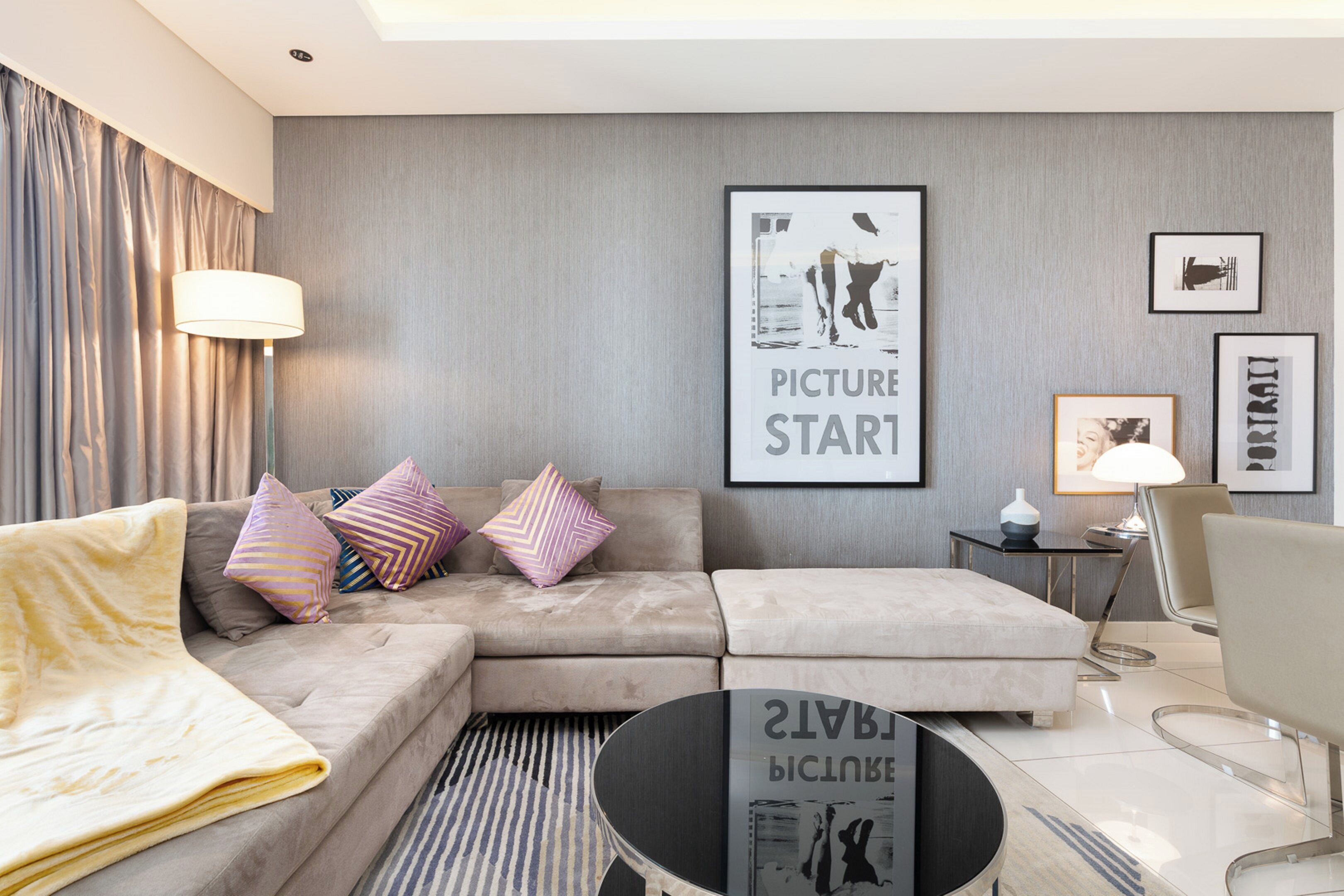 A modern living room with a sofa, ottoman, and a coffee table. There are framed pictures on the wall and a lamp on a side table.