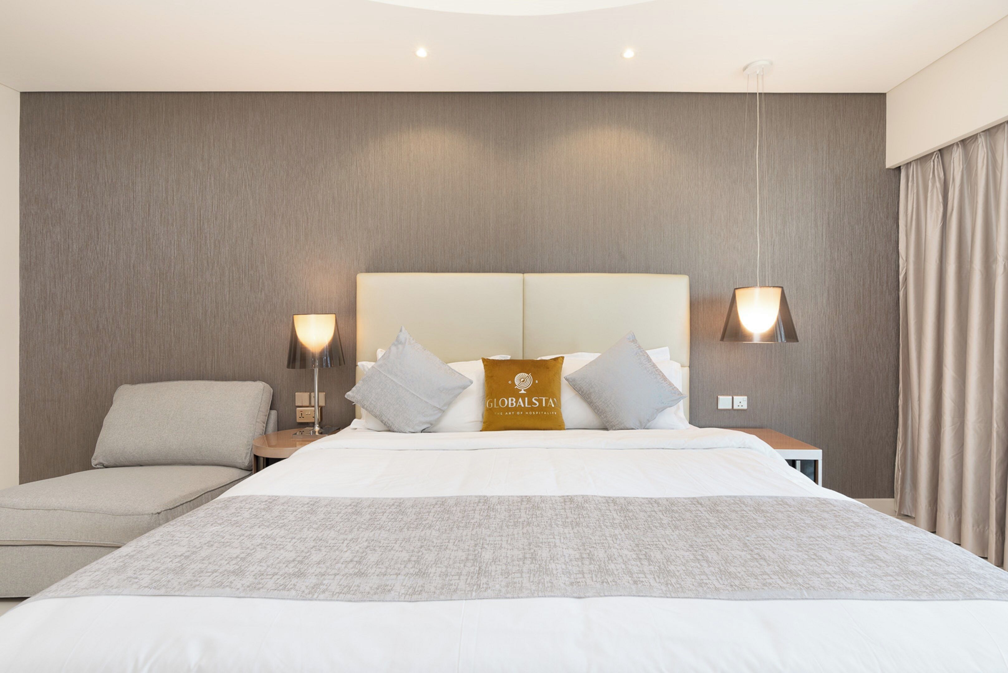A modern hotel room with a large bed, two bedside tables, a sofa, and a headboard with a logo.