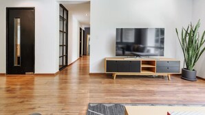 Ein modernes Wohnzimmer mit Holzboden, einem Fernseher auf einem Holz-TV-Möbel und einer Zimmerpflanze, sowie einer Tür zu einem weiteren Raum.