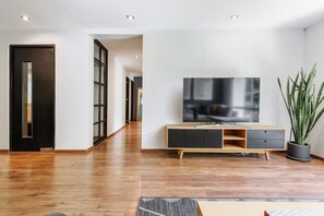 Ein modernes Wohnzimmer mit Holzboden, einem Fernseher auf einem Holz-TV-Möbel und einer Zimmerpflanze, sowie einer Tür zu einem weiteren Raum.