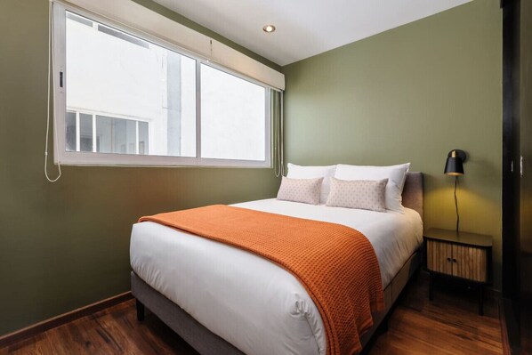 Ein Schlafzimmer mit großem Fenster, einem Bett mit weißer Bettwäsche und einer orangefarbenen Decke, einem Nachttisch mit Lampe und Holzboden.