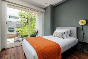 Ein Schlafzimmer mit Bett, Nachttisch, Stuhl, Fenster mit Blick auf Grünflächen und ein Gebäude sowie einer Deckenleuchte.