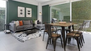 A modern living room with a grey sofa, a dining table with chairs, and a large window overlooking a green area. - Stylish 2-Bdr Haven with Relaxing Balcony (Ciudad de México)