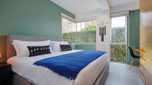 A modern bedroom with a large bed, a nightstand, a chair, and a view of greenery outside. - Stylish 2-Bdr Haven with Relaxing Balcony (Ciudad de México)