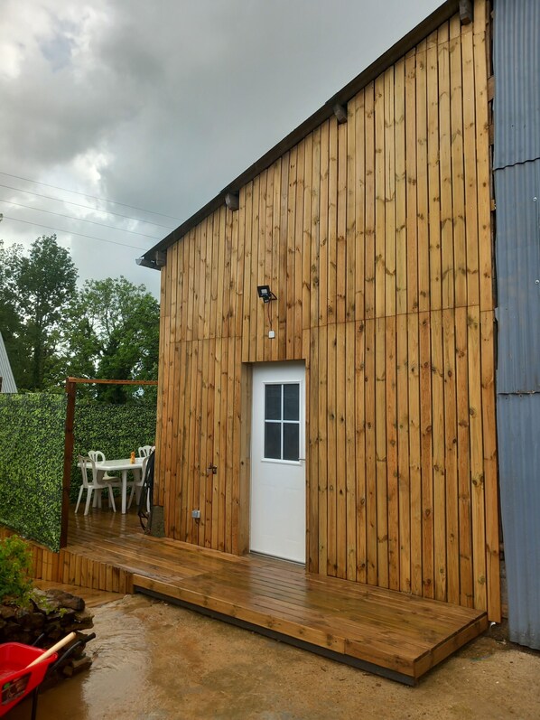 Un bâtiment en bois avec une porte blanche et une petite fenêtre, un espace extérieur aménagé avec des sièges et un pont en bois.
