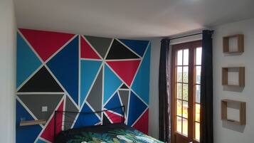 Une chambre avec un mur orné d’un motif géométrique, un lit recouvert d’une housse à motifs de feuilles, une fenêtre avec des rideaux et une porte.