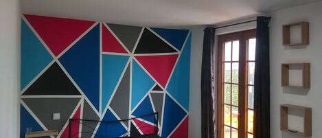 Ein Schlafzimmer mit Wand in geometrischem Muster, einem Bett mit blattbedecktem Überwurf, einem Fenster mit Vorhängen und einer Tür.