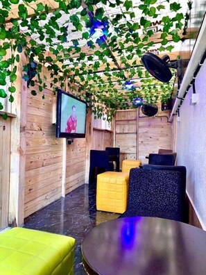 A cozy room with wooden walls, a flat-screen TV mounted on the wall, and a ceiling covered with green foliage. There are yellow and black cushioned seating areas and a round table in the foreground. - Vintage Classic Suites (Lekki)