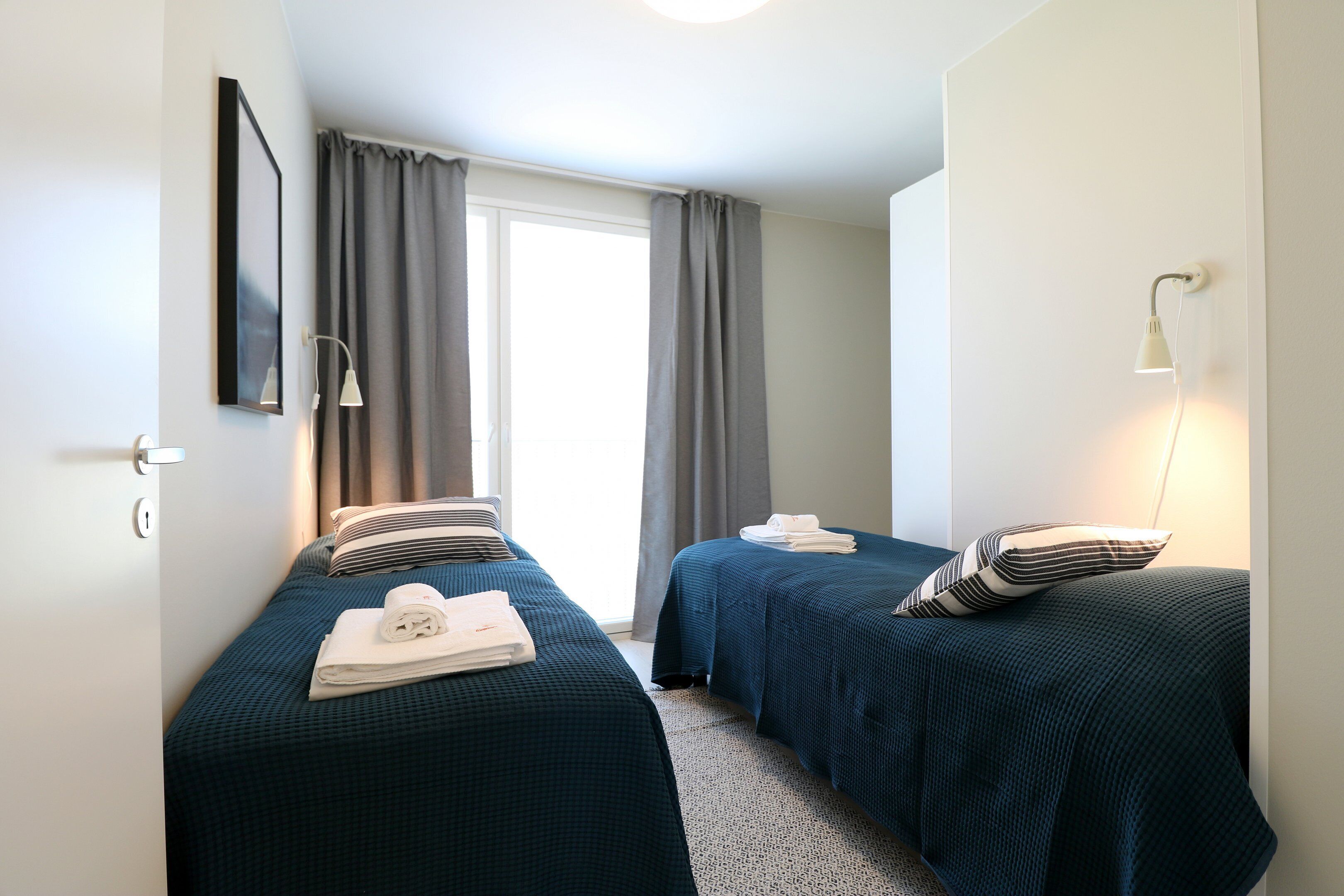 Una camera d'albergo con due letti, una grande finestra con tende, una lampada fissata al muro e un quadro incorniciato.