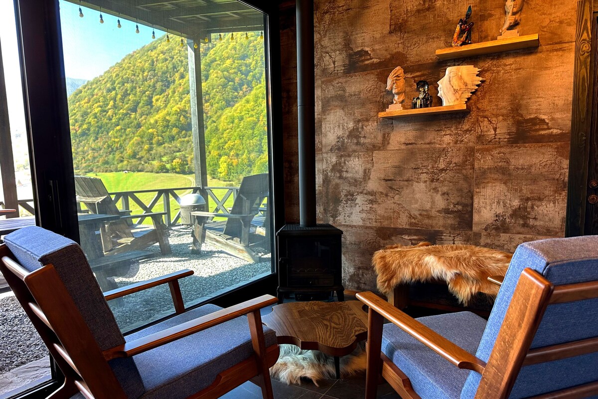 A cozy room with a large window overlooking a deck and a forested hill. There are two wooden chairs with blue cushions, a small fur-covered ottoman, and a fireplace.