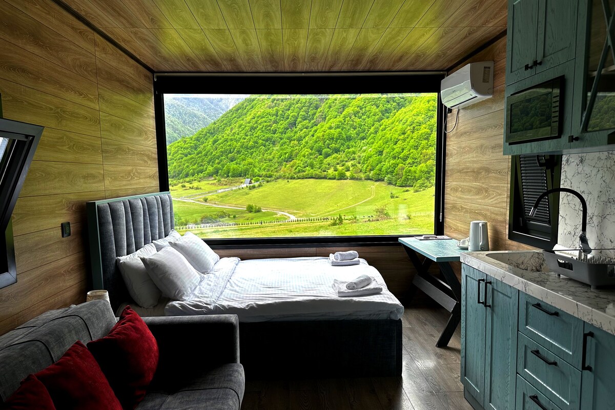 A bedroom with a large window, a bed, a sofa, and a small table with a kettle.
