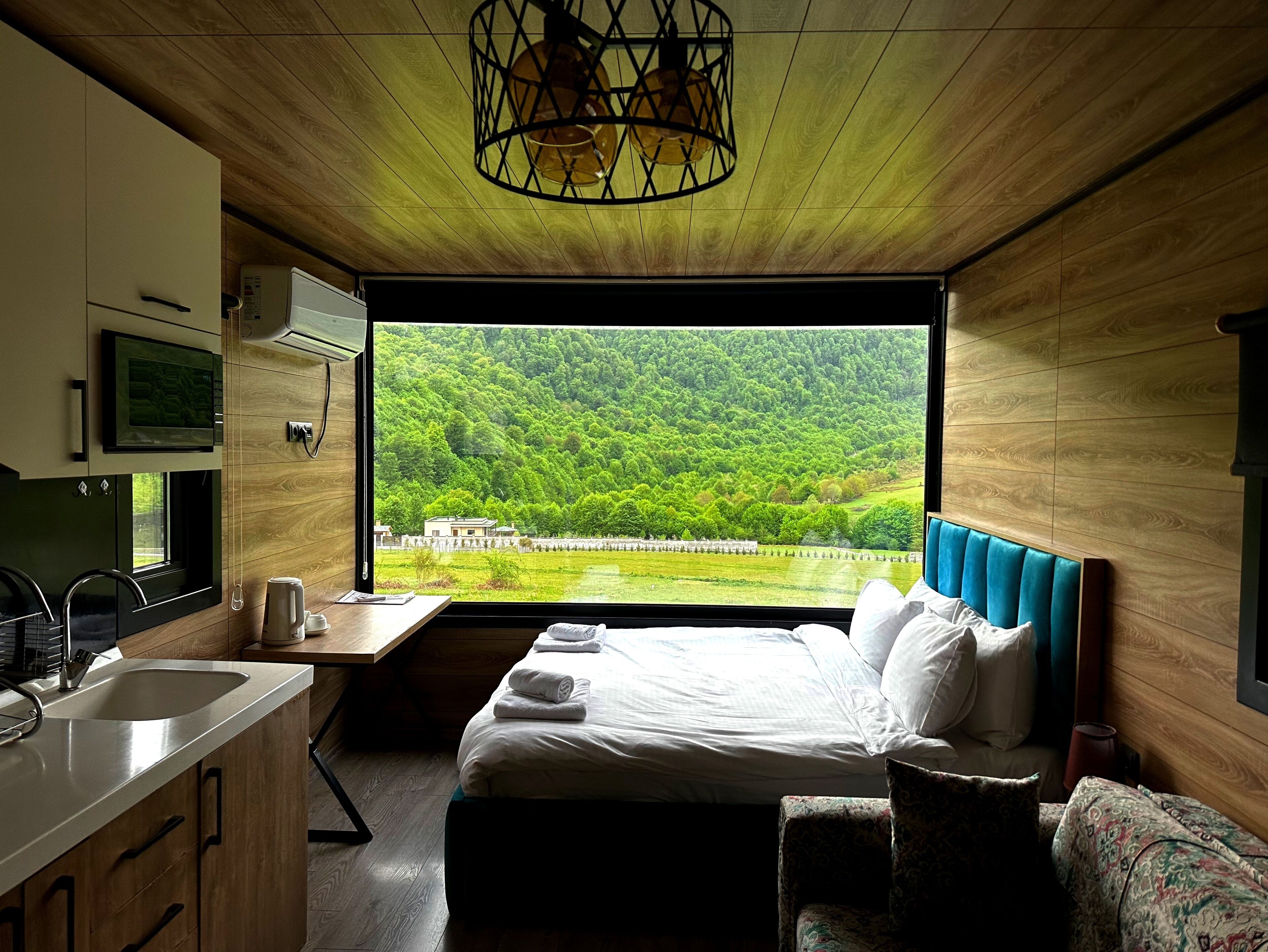 A modern bedroom with a large window, a bed, a desk, and a kitchen area.