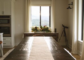 A dining room with a wooden table, a window with a view of a forest, a telescope, and a cabinet with appliances.