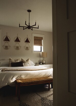 A bedroom with a bed, a bedside table with a lamp, a window with blinds, and a wall-mounted light fixture.