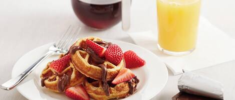 A plate of waffles with strawberries and chocolate sauce, a glass of orange juice, and a chocolate bar on a table.