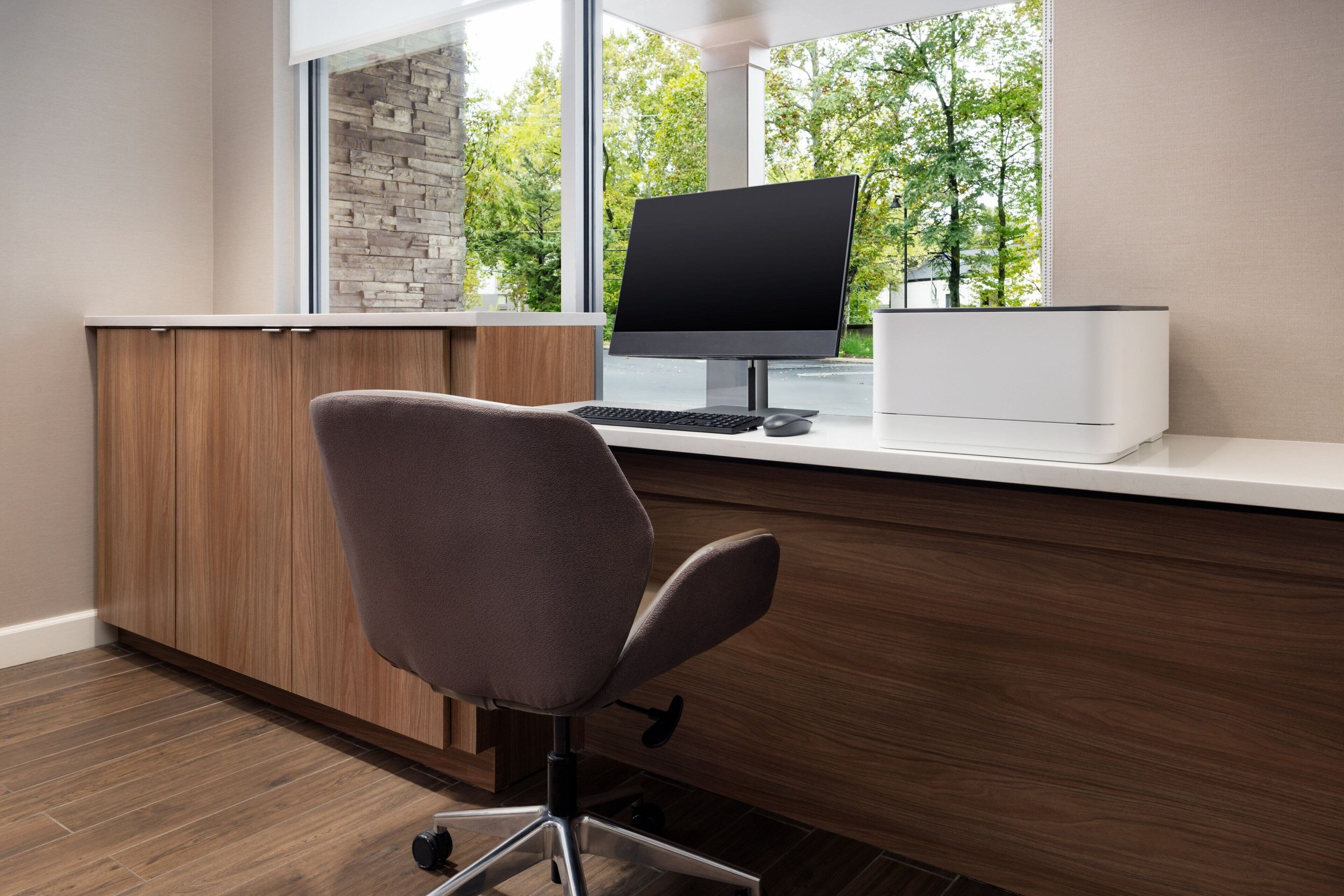 A modern office desk with a computer, a chair, and a view of trees through a window.