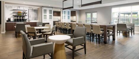 A spacious dining area with multiple tables and chairs, a kitchen in the background, and large windows providing natural light.
