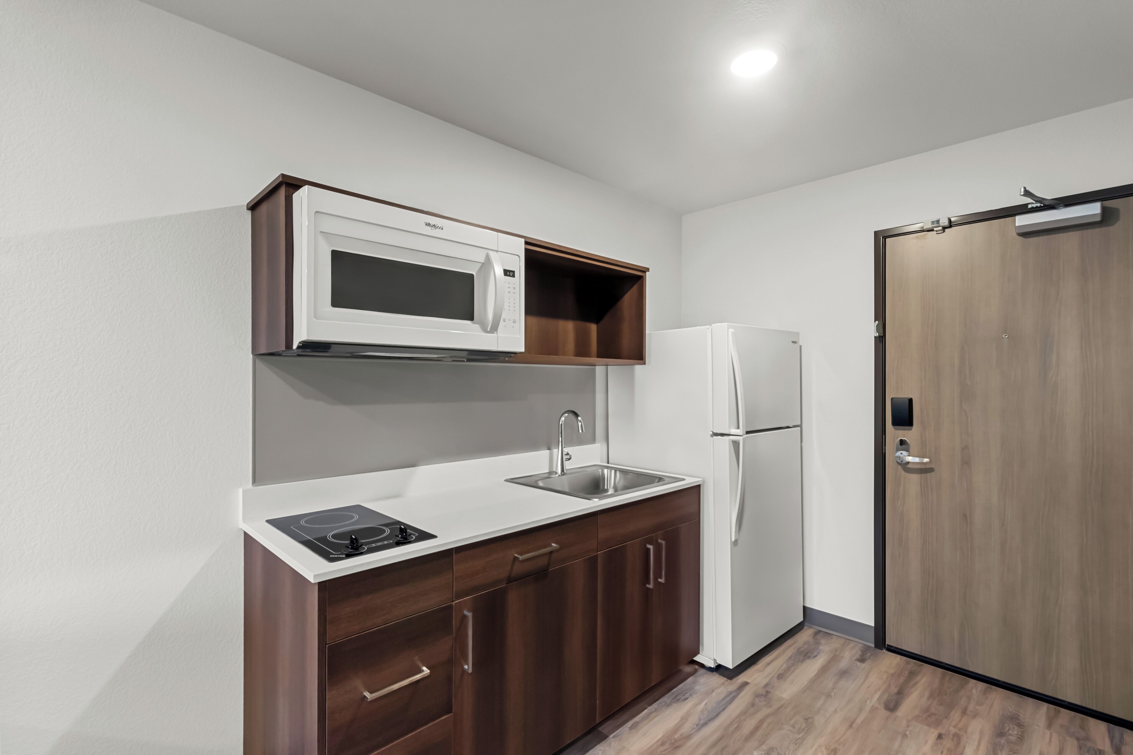 A compact kitchen with a microwave, sink, and refrigerator, wooden cabinets, and a door with a handle.
