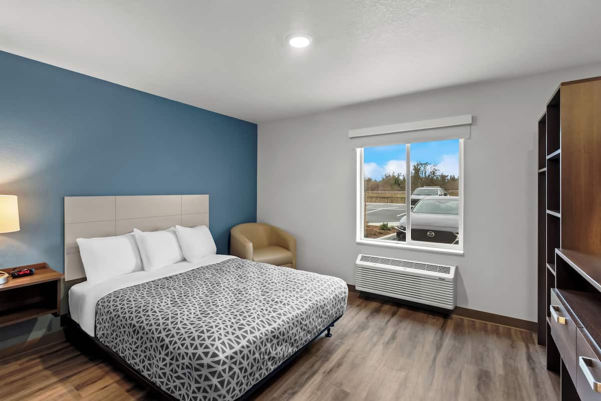 A modern hotel room with a large bed, a chair, a window with a view of a parking lot, and built-in storage.