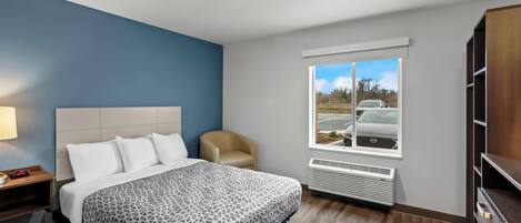A modern hotel room with a large bed, a chair, a window with a view of a parking lot, and built-in storage.