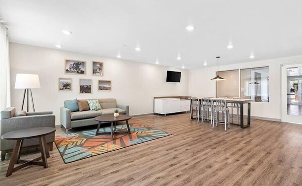 A modern living room with a sofa, a coffee table, a dining area with chairs, a TV mounted on the wall, and artwork on the wall. - WoodSpring Suites Beaverton - Portland West (Beaverton)