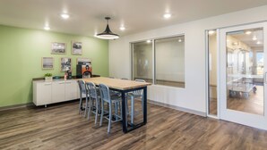 A modern dining area with a wooden table and chairs, a white cabinet with plants, and a large window. - WoodSpring Suites Beaverton - Portland West (Beaverton)