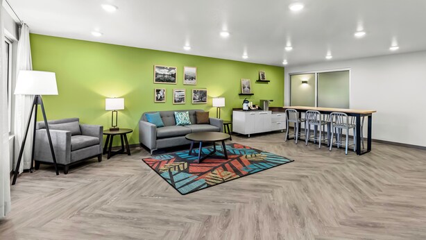 A modern living room with a green accent wall, a sofa, a coffee table, and a dining area with chairs. - WoodSpring Suites Beaverton - Portland West (Beaverton)
