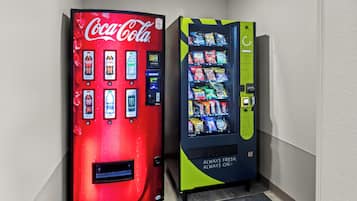 Two vending machines, one Coca-Cola branded and one with a green and yellow design, in a hallway.