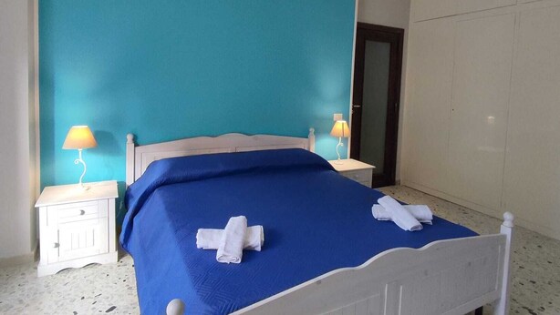 Un dormitorio con una colcha azul, dos lámparas de noche, una mesita de noche blanca y una puerta al fondo.