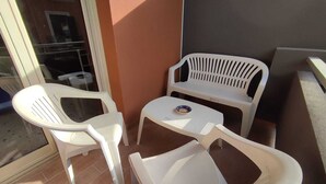 Un pequeño espacio al aire libre con dos sillas de plástico blancas y una mesa, un vidrio y suelo de baldosas.