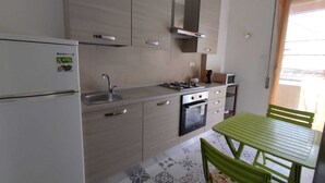 Cocina compacta con refrigerador, estufa, lavamanos y armarios. Hay una mesa de comedor con sillas y un ventanal que proporciona luz natural.
