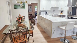 Una cocina moderna con armarios blancos, una isla de mármol y una zona de comedor con mesa de vidrio y sillas de metal.