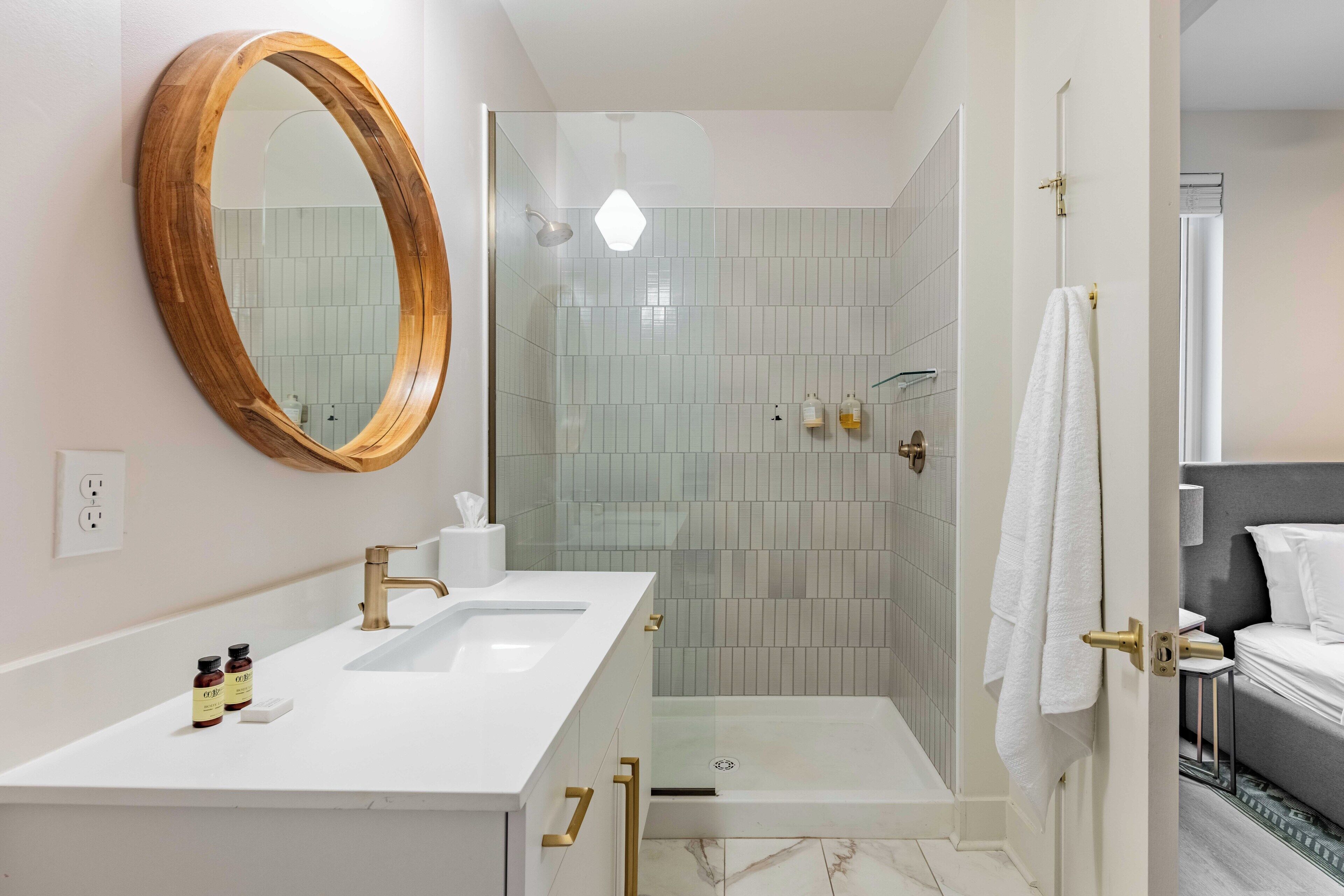 Un baño moderno con un tocador blanco, un espejo grande, una ducha con puertas de vidrio y una toalla colgada en la puerta.