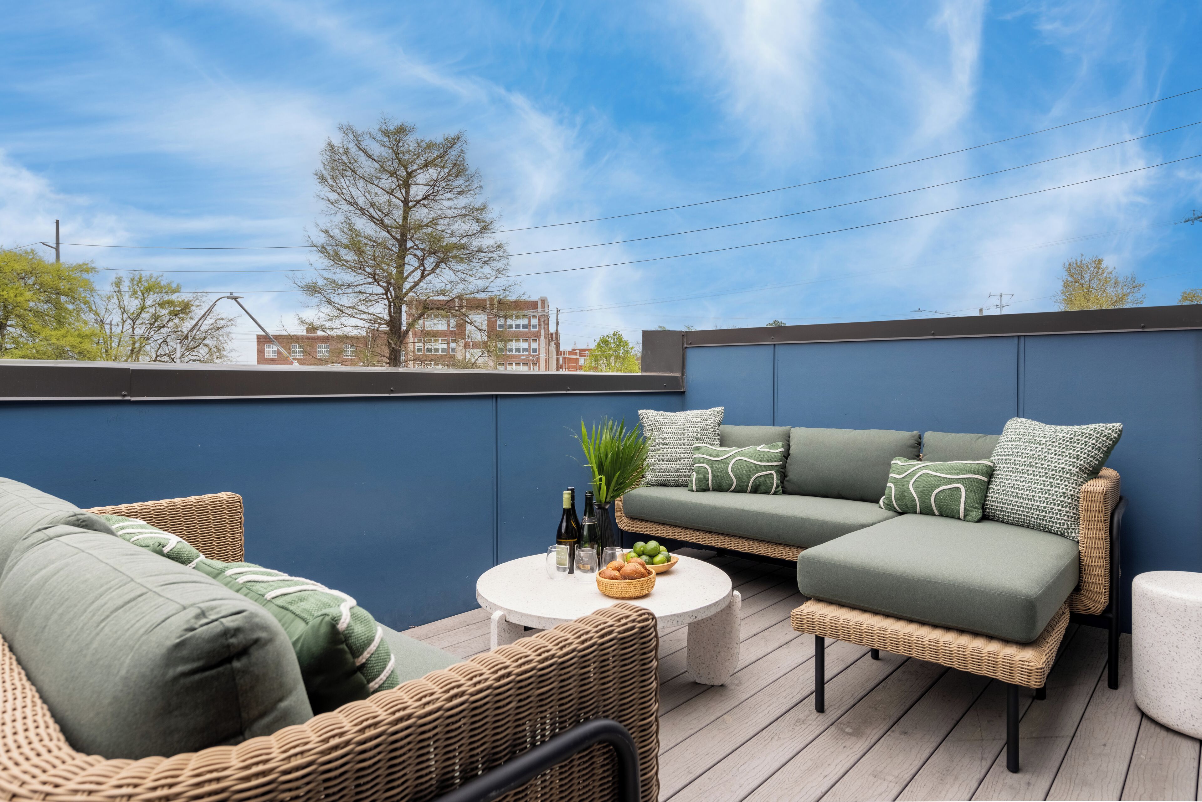 A rooftop terrace with wicker furniture, a green sofa, and a round table with bottles and a bowl of fruit.