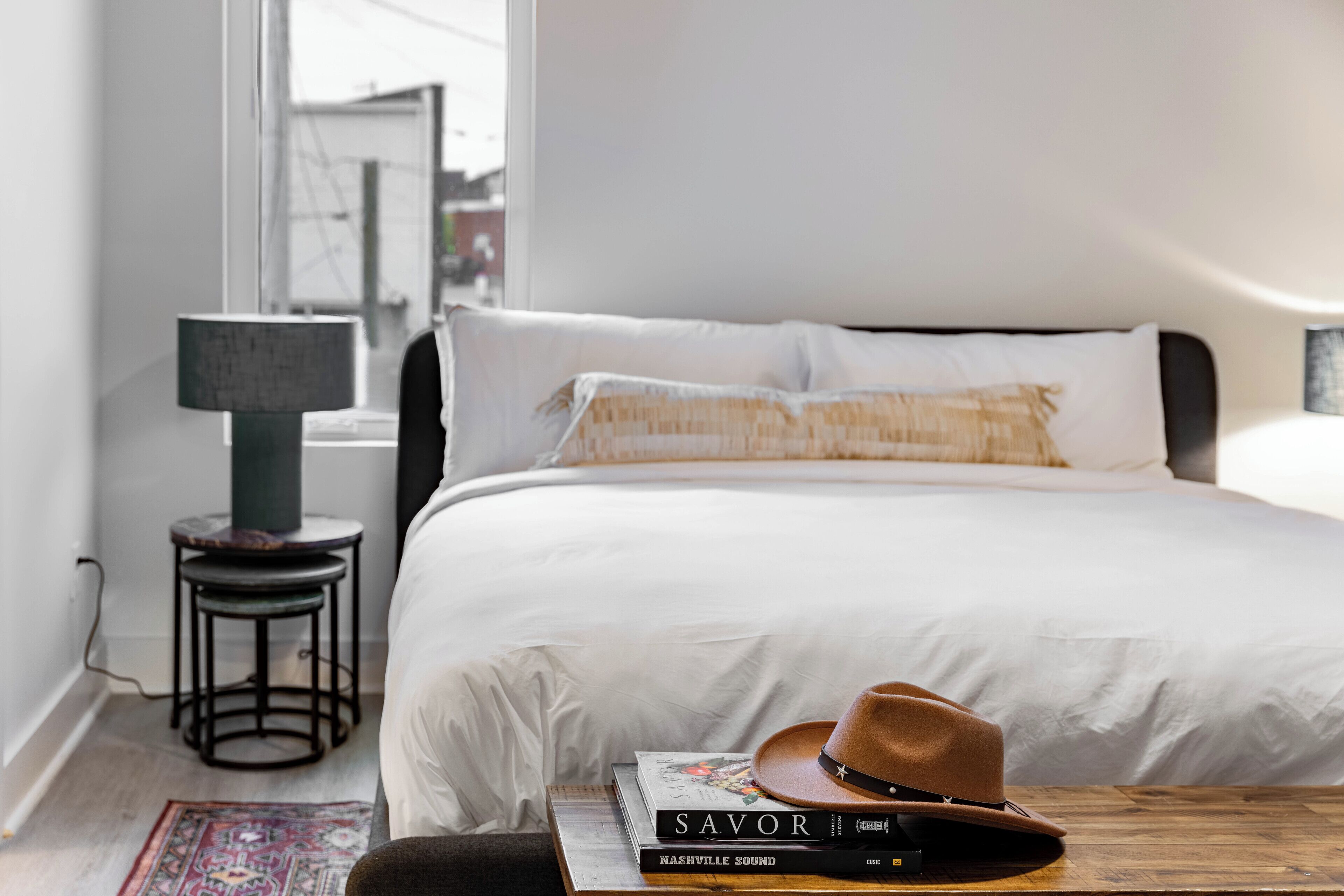 A neatly made bed with white linens, a small round table with a lamp, and a few books and a hat on the bed.