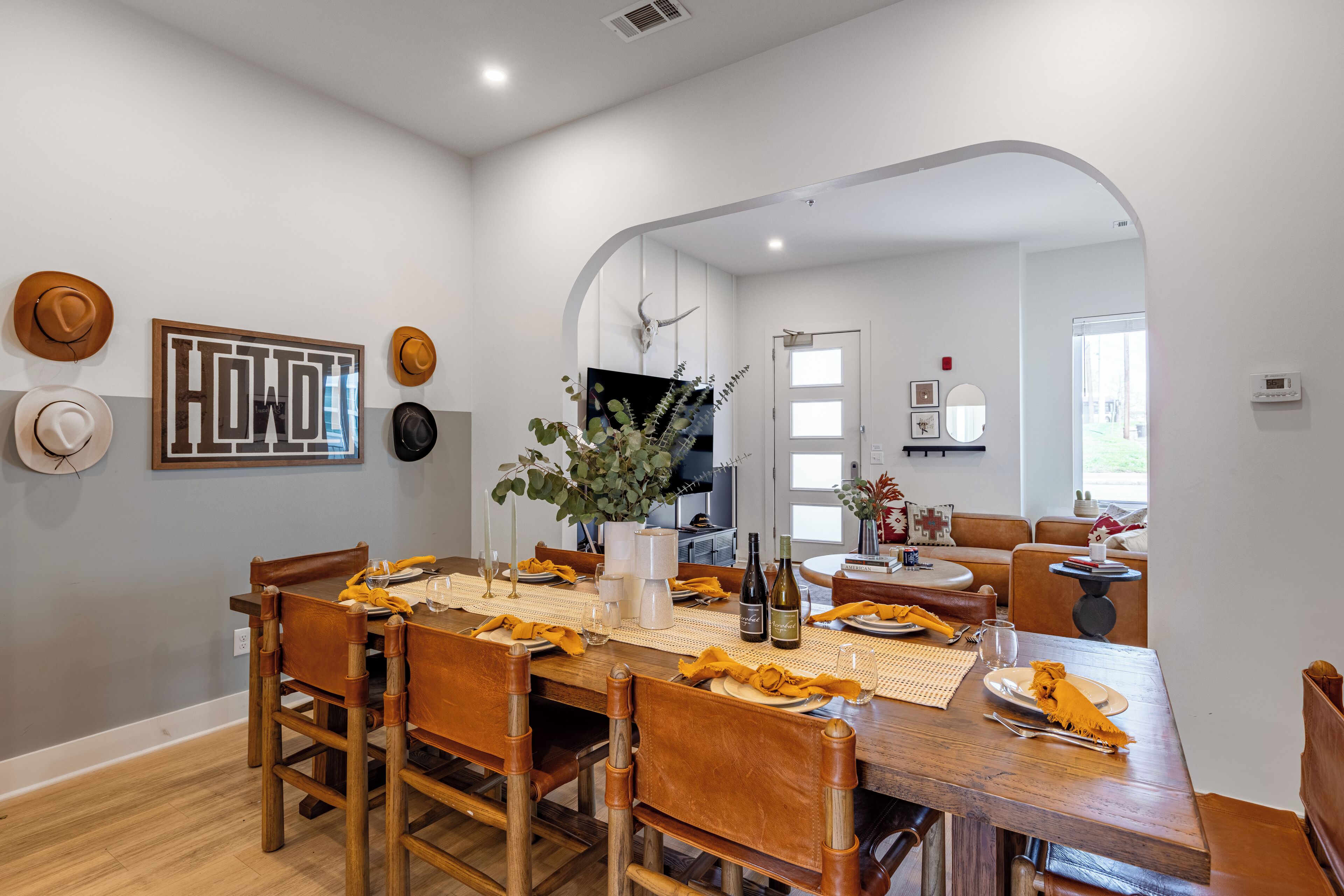 A dining area with a wooden table set for four, surrounded by chairs. There are hats on the wall, a large framed word art, and a plant on the table.