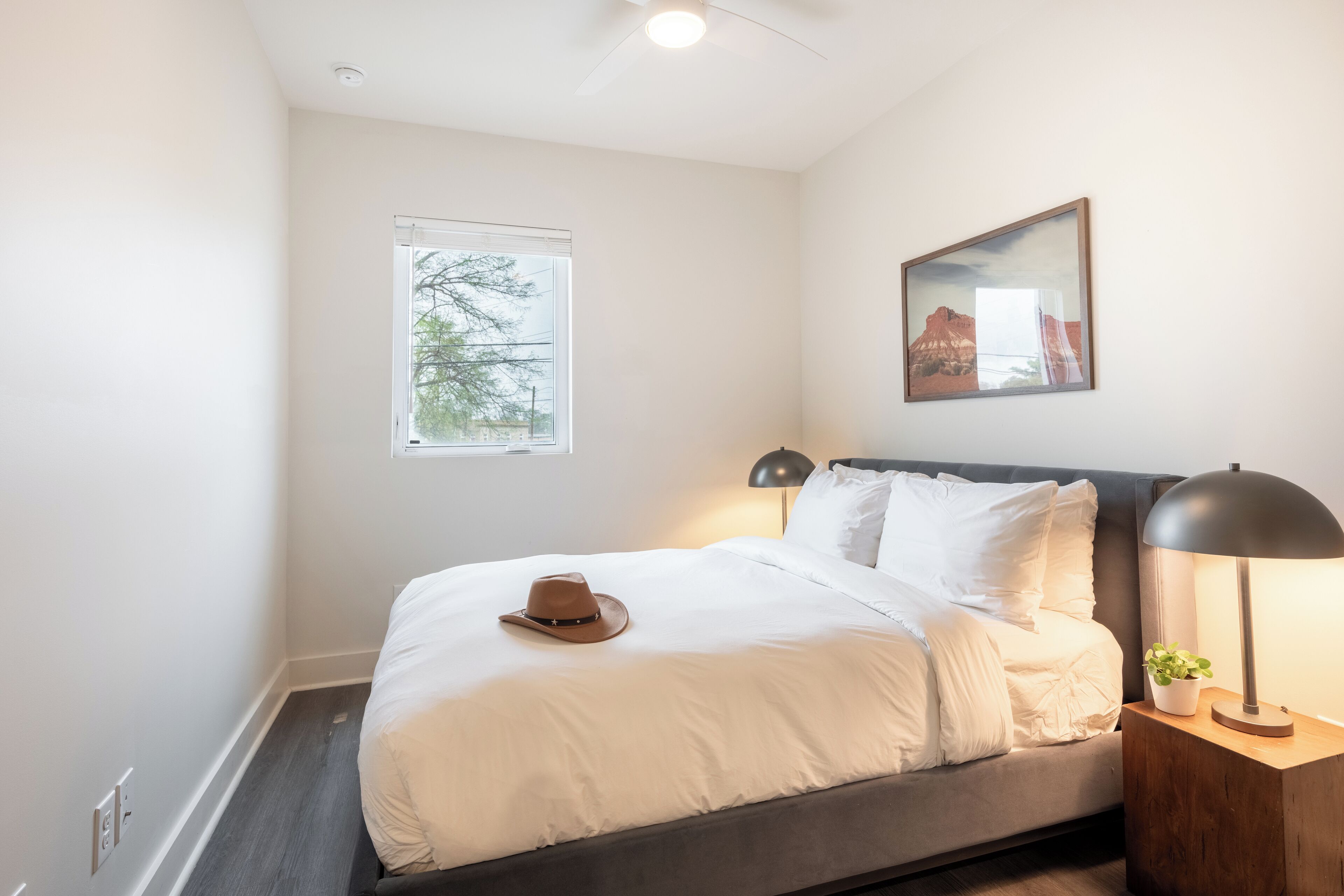 A neatly made bed with white linens, a brown hat, and a small potted plant beside a bedside table with a lamp.