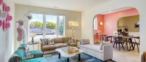 A spacious living area with a large window, a dining area, and a kitchen visible through an archway.