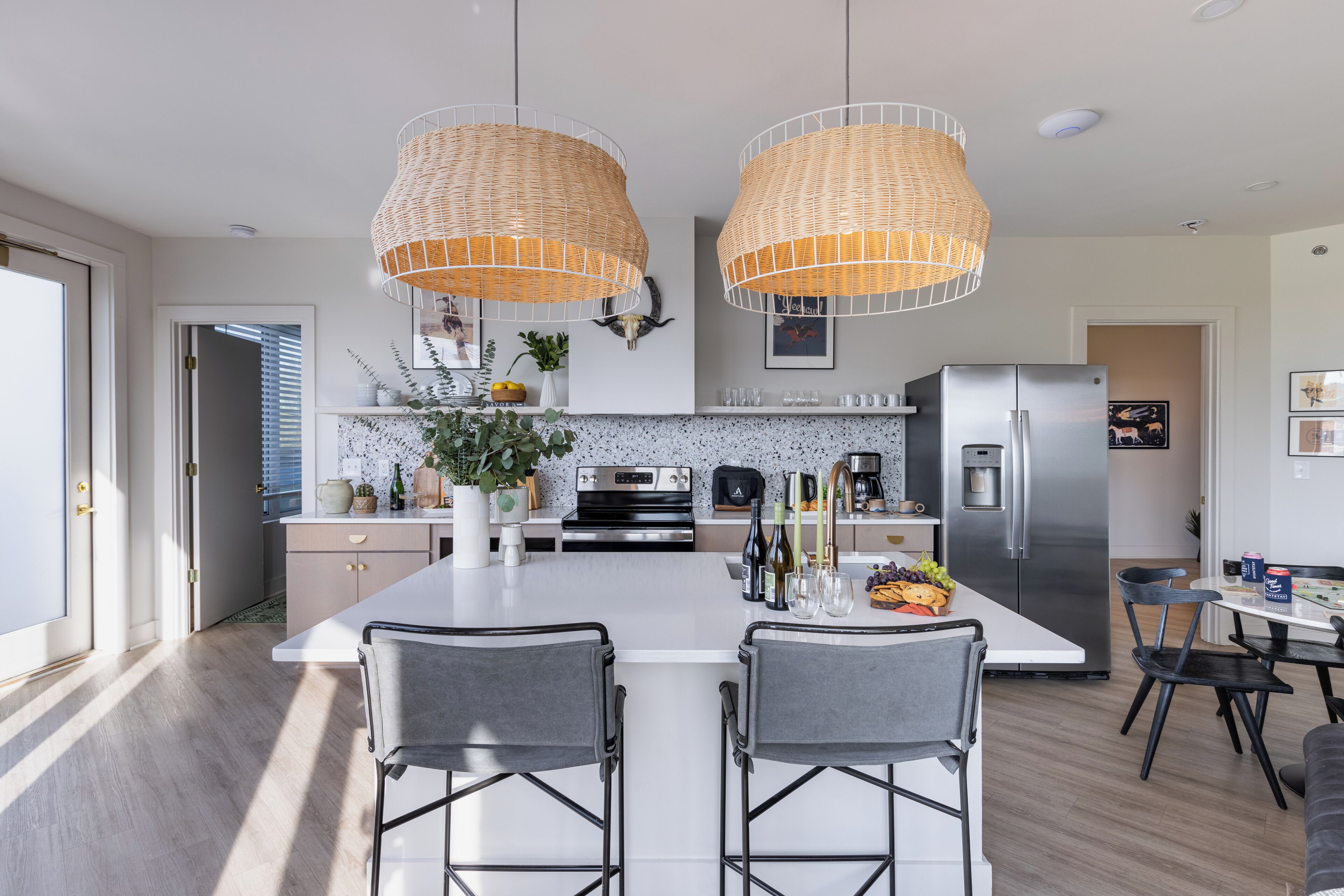 A modern kitchen with a central island, two bar stools, a stainless steel refrigerator, and hanging wicker pendant lights.