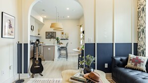 Una sala de estar moderna con un sofá de cuero negro, una mesa de centro con libros y un sombrero, una guitarra, y un comedor con una araña.