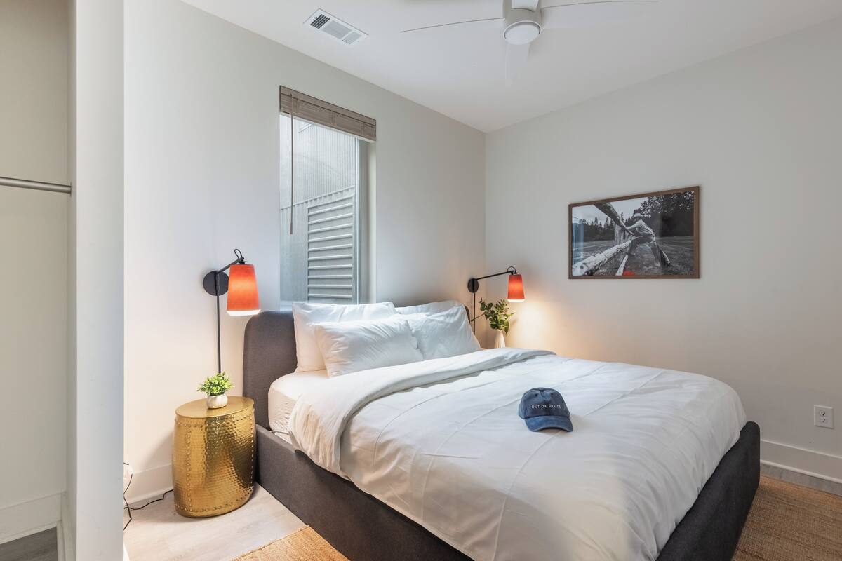 A bedroom with a bed, two bedside lamps, a window with blinds, a wall-mounted picture, and a ceiling fan.