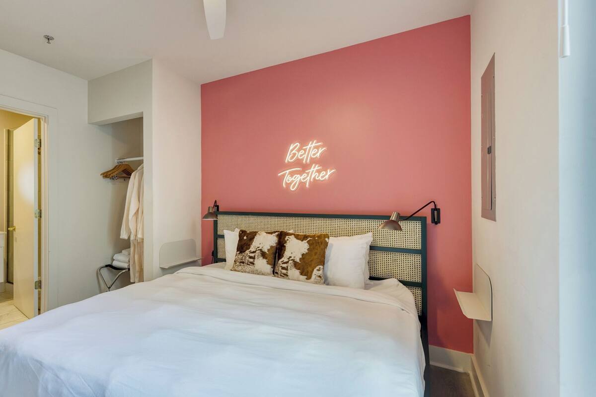 A bedroom with a bed, a bedside lamp, a wall with a neon sign, a wardrobe, and a bathroom visible through an open door.