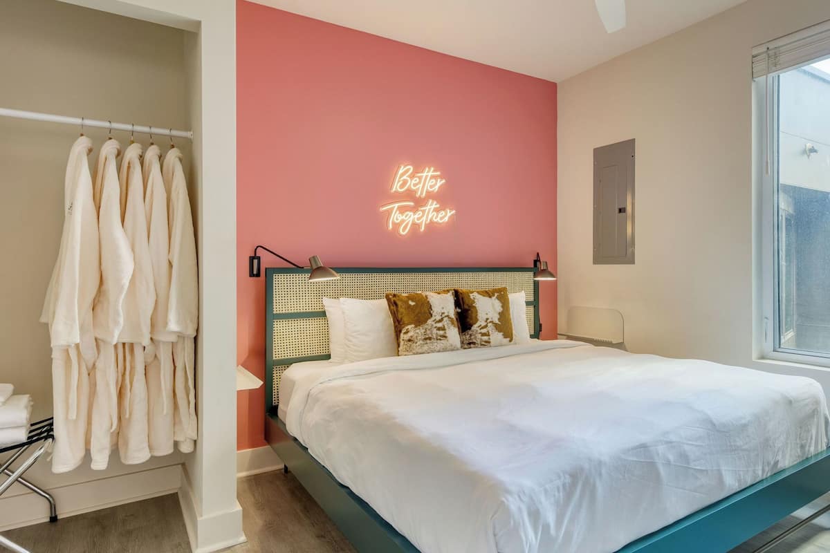 Ein Schlafzimmer mit einem Bett, einer Neonreklame, einem Fenster und einem Kleiderständer.