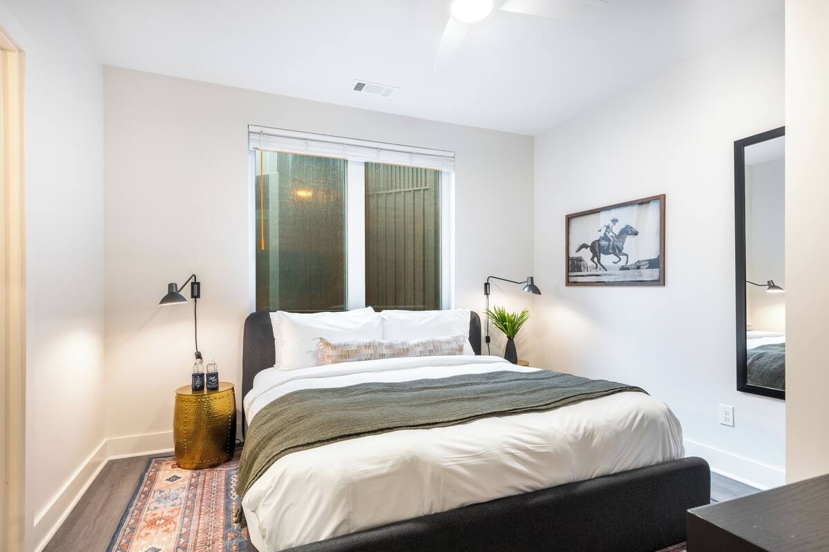 A bedroom with a bed, bedside lamps, a window, a framed picture, and a small plant.