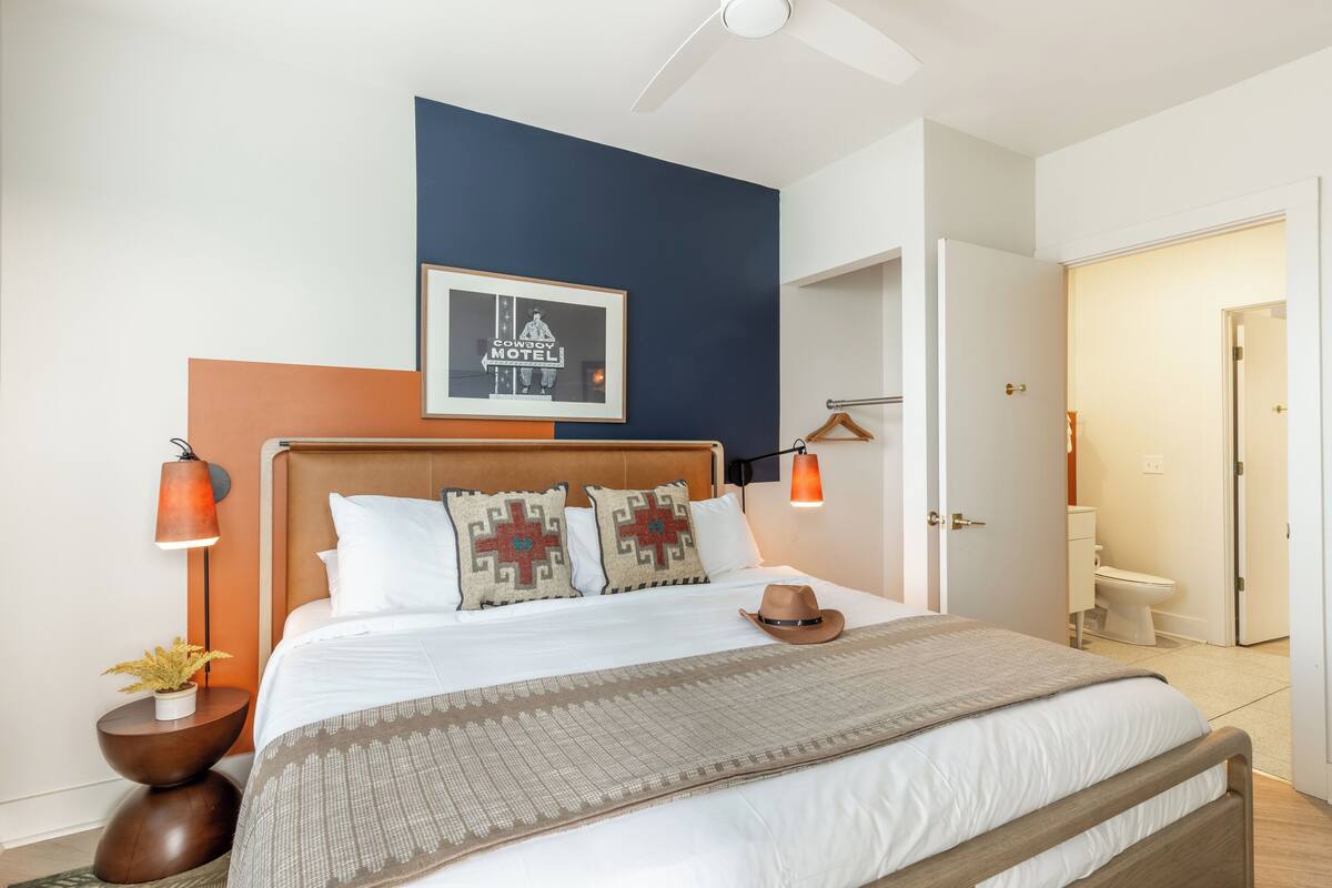 A neatly arranged bedroom with a bed, pillows, a hat, a framed picture, and a nightstand with a lamp.