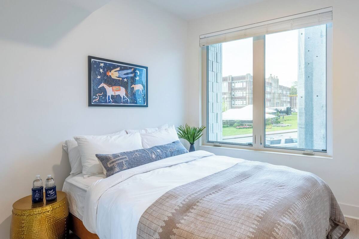 A bedroom with a bed, pillows, a nightstand with bottles, a window with a view of buildings, and a wall-mounted picture.