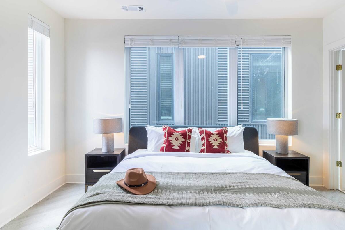A bedroom with a bed, two bedside tables, a window with blinds, and a door.