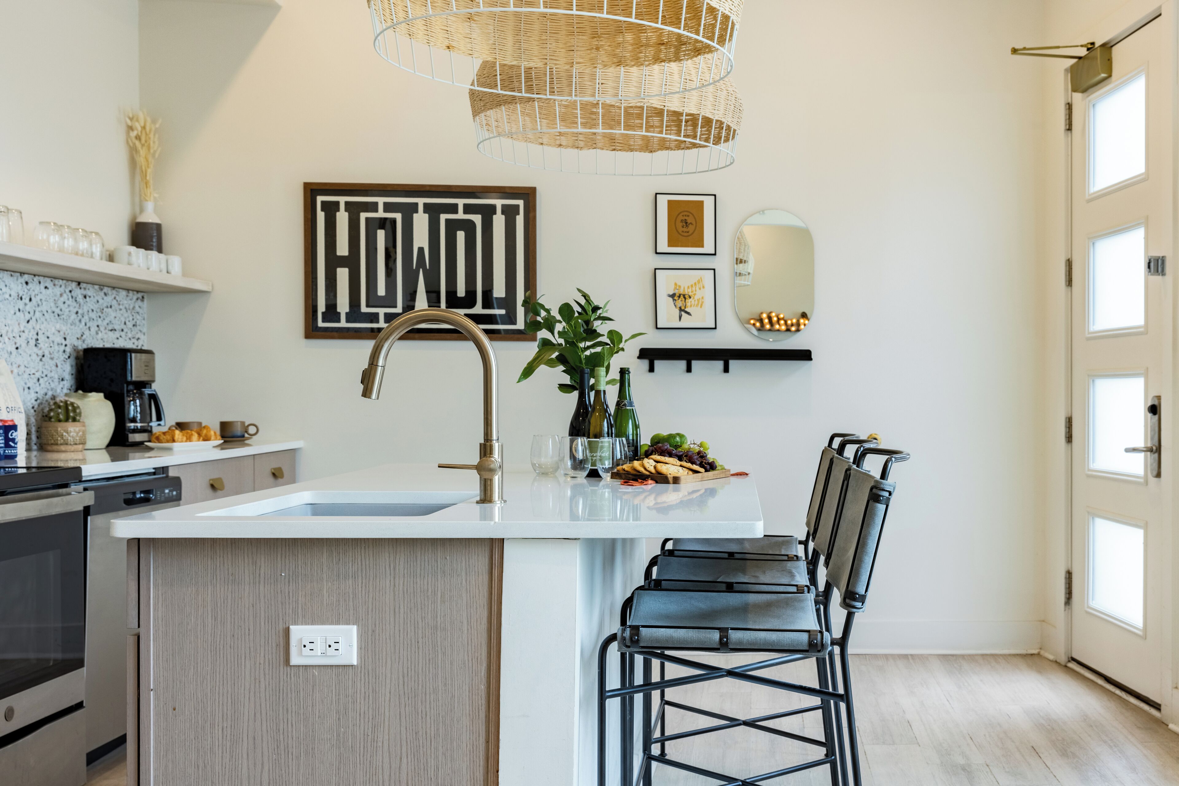 A modern kitchen with a central island, black bar stools, a hanging light fixture, and a 'HOWL' sign on the wall.